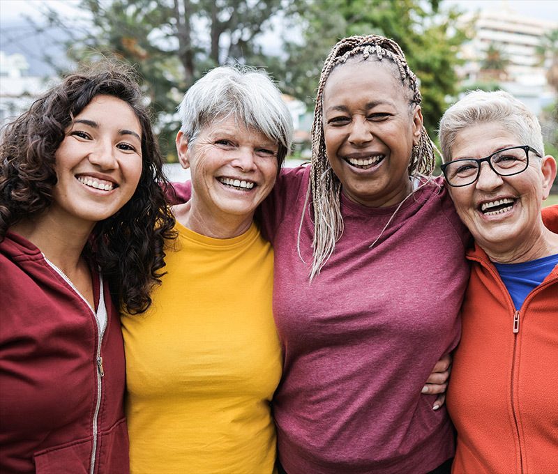 Four smiling happy women of varying ages and races with arms around each other in outside setting.