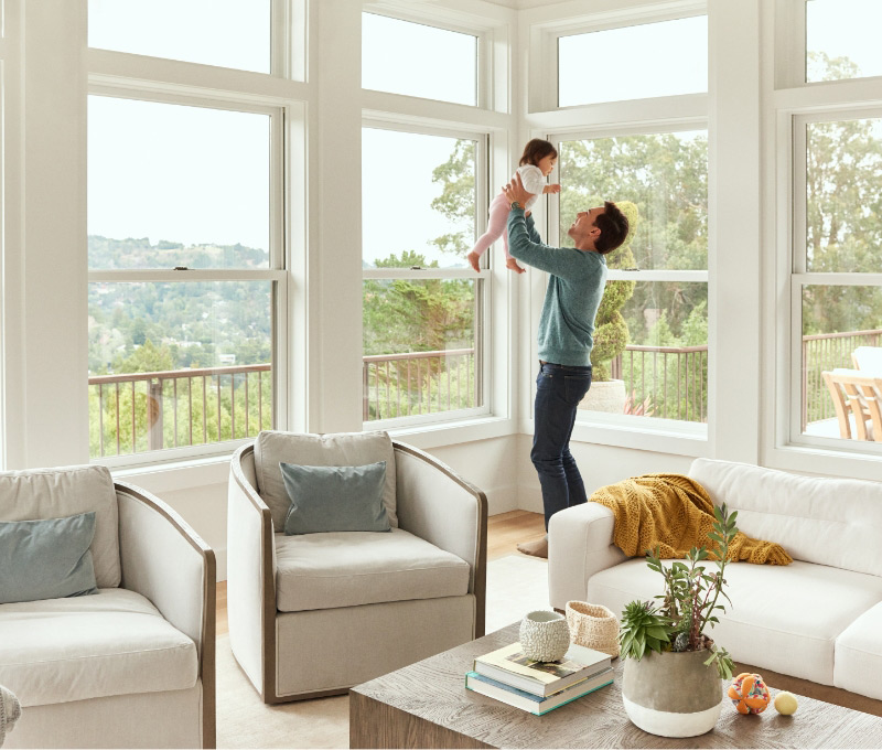 Image of a father holding up a baby girl while laughing in well lit living room setting with view of treetops outside.