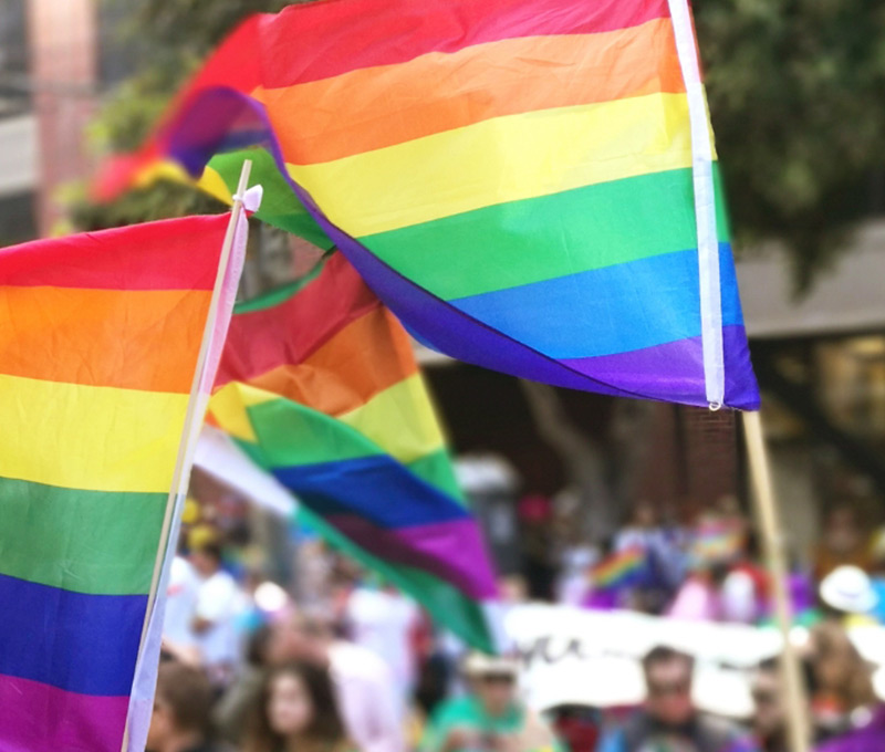 Several rainbow flags representing LGBTQ+ rights waving in foreground of a Pride parade.